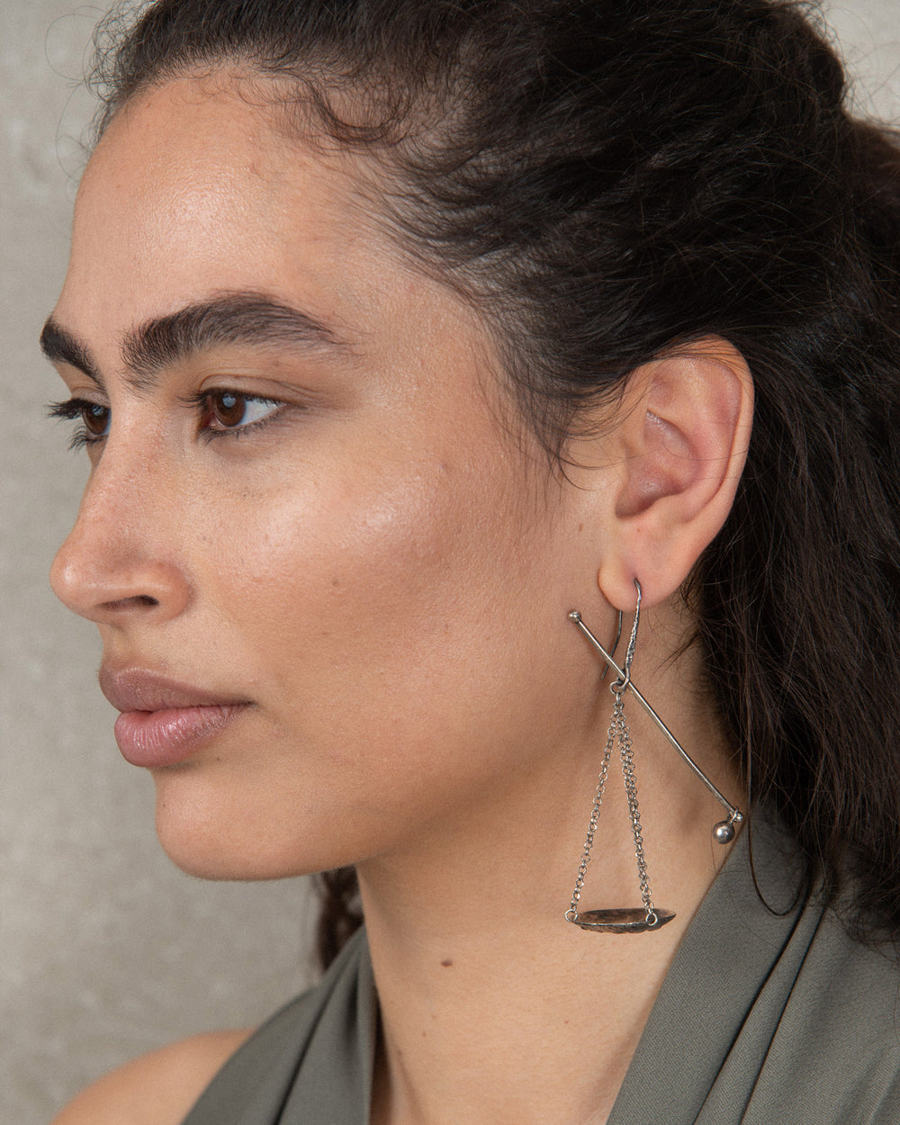 A single silver dangling earring featuring a scale or balance weight. Worn by a sophisticated woman.