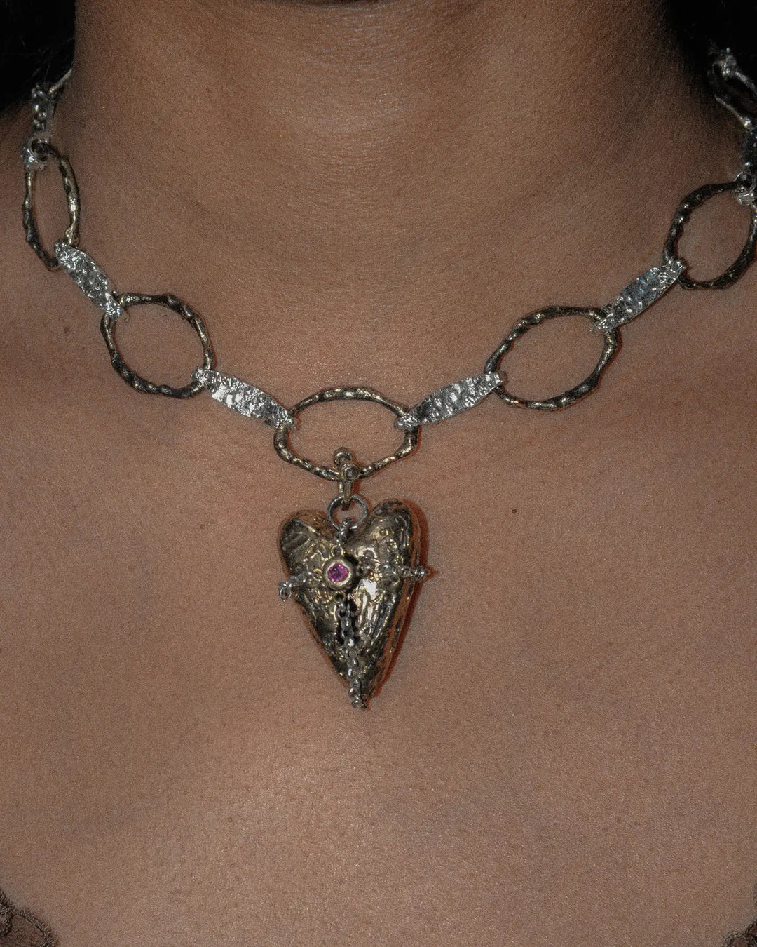 Close-up of a bronze heart-shaped charm with textured finish, attached to a silver chain necklace. The detailed design highlights the intricate craftsmanship of the heart pendant and the chain's delicate links.
