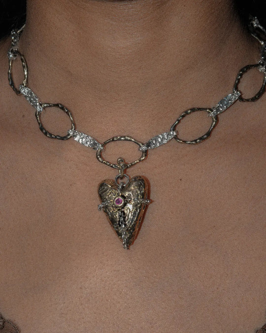 Close-up of a bronze heart-shaped charm with textured finish, attached to a silver chain necklace. The detailed design highlights the intricate craftsmanship of the heart pendant and the chain's delicate links.