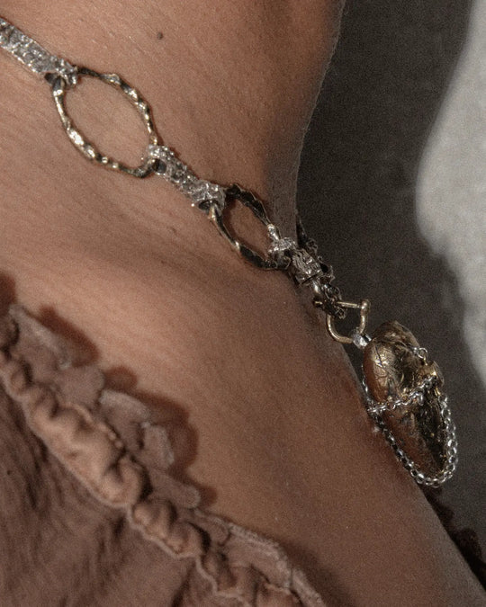 Side view of a chain necklace featuring a bronze heart-shaped charm with textured finish, highlighting the three-dimensional design and detailed craftsmanship of the pendant.