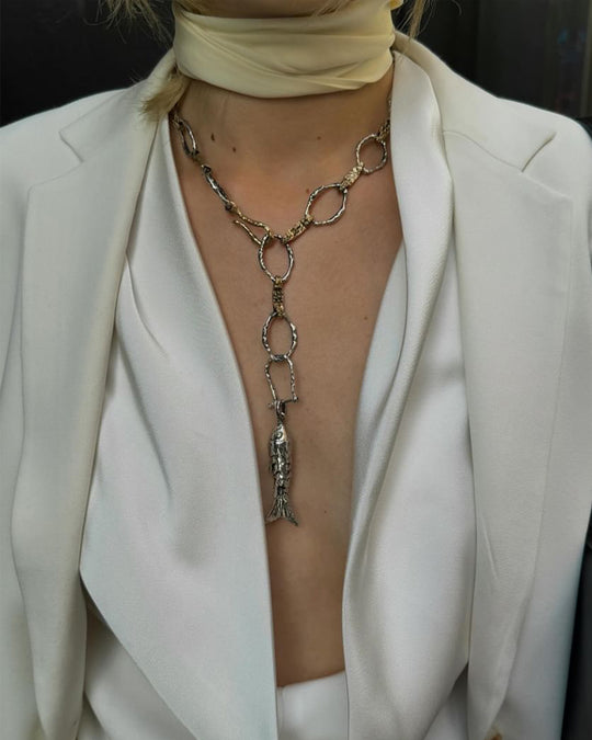 Iconic Christie Tyler wearing a white blazer with a long, intricate necklace featuring a fish pendant. Giving lux chic vibes.