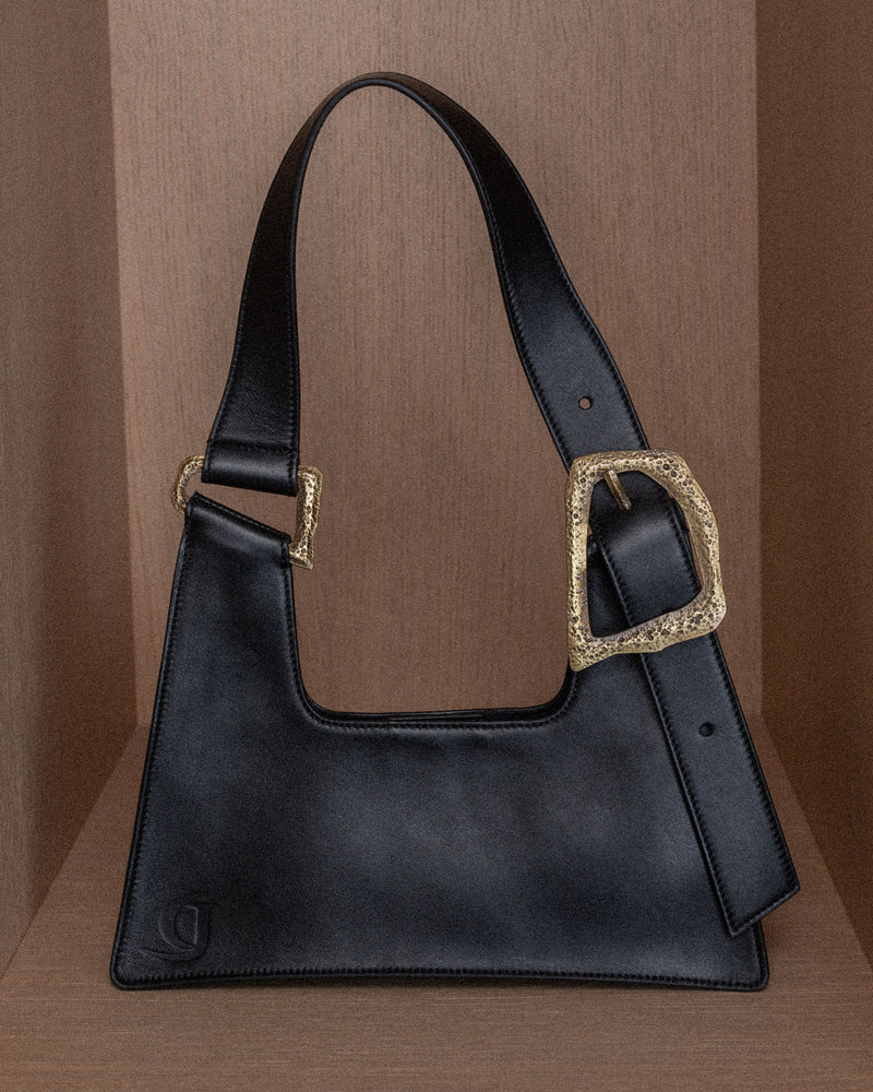 A black handbag with bronze metal hardware and an adjustable strap, displayed against a wooden background.