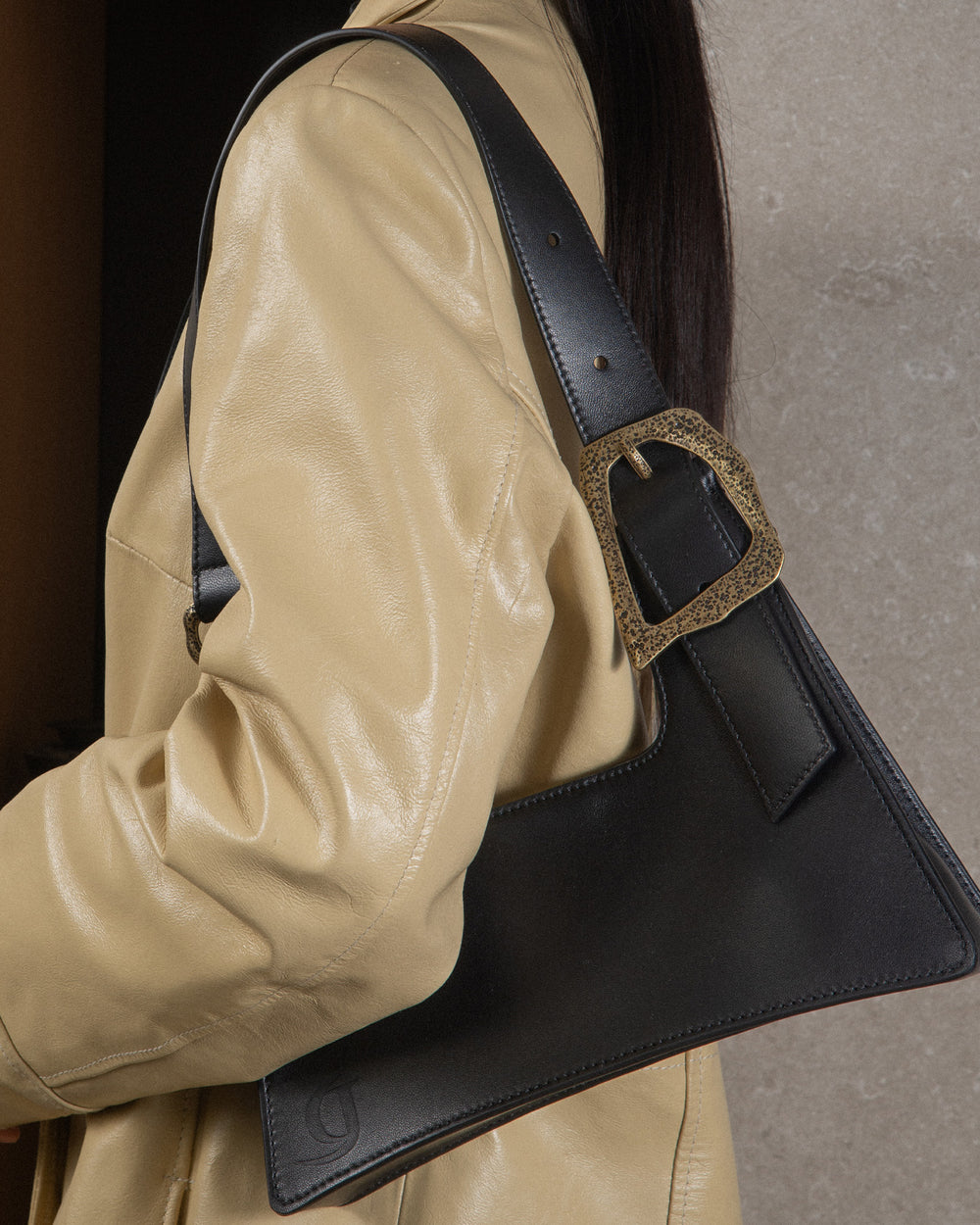 A woman holding a sleek black upcycled leather bag with detailed bronze accents. The bag features visible topstitches that showcase luxurious craftsmanship, highlighting its high-quality construction. She is dressed in a butter yellow trench coat, creating a vibrant and sophisticated look that emphasizes elegance and sustainable style.