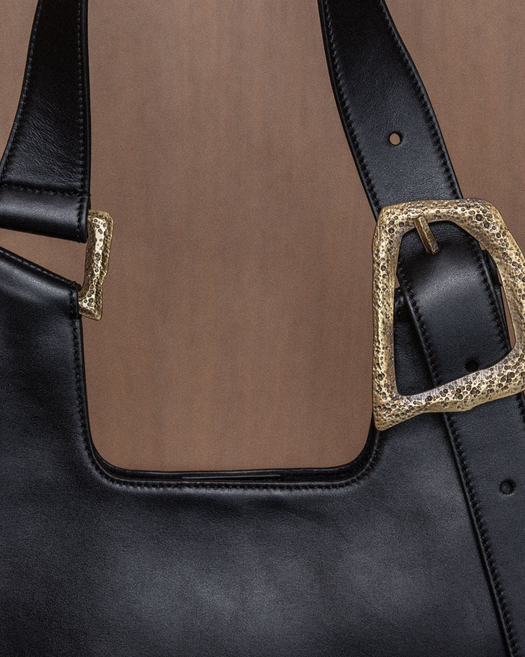 A still life composition featuring a luxurious black upcycled leather bag with intricate bronze details, set against a warm wooden background. The focus highlights the rich texture and high-quality craftsmanship of the leather, with visible topstitches that emphasize the artisanal design and refined bronze accents, creating an elegant and timeless display.