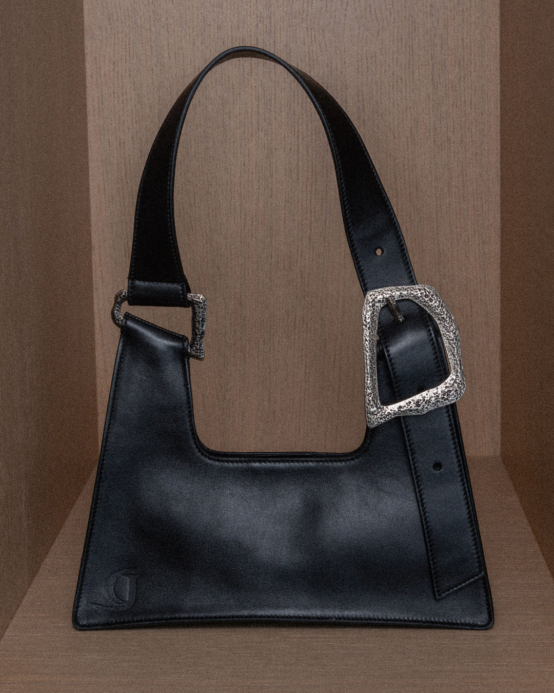 A still life composition featuring a sleek black upcycled leather bag with silver details, set against a warm wooden background. The focus highlights the smooth texture and high-quality craftsmanship of the leather, complemented by the elegant silver accents that add a modern touch. The scene emphasizes the bag's luxurious design and artisanal craftsmanship.