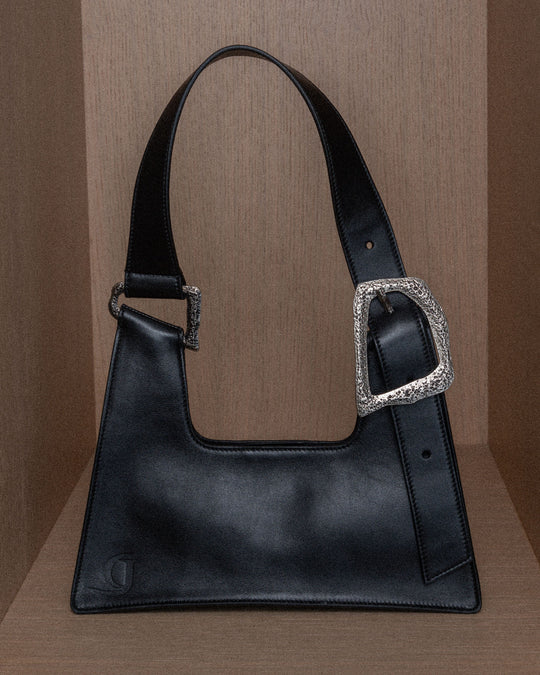 A still life composition featuring a sleek black upcycled leather bag with silver details, set against a warm wooden background. The focus highlights the smooth texture and high-quality craftsmanship of the leather, complemented by the elegant silver accents that add a modern touch. The scene emphasizes the bag's luxurious design and artisanal craftsmanship.
