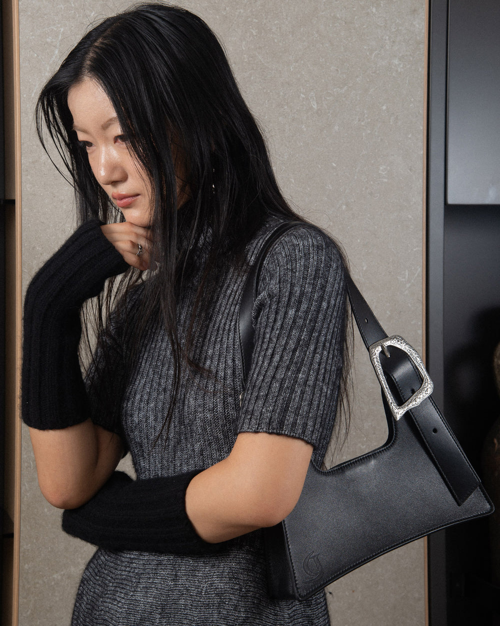 A woman wearing a minimalist grey knitwear dress, carrying a sleek black upcycled leather bag with silver details on her shoulder.