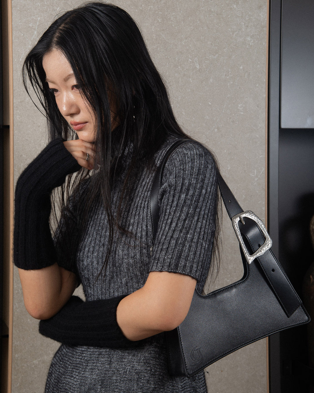 A woman wearing a minimalist grey knitwear dress, carrying a sleek black upcycled leather bag with silver details on her shoulder.