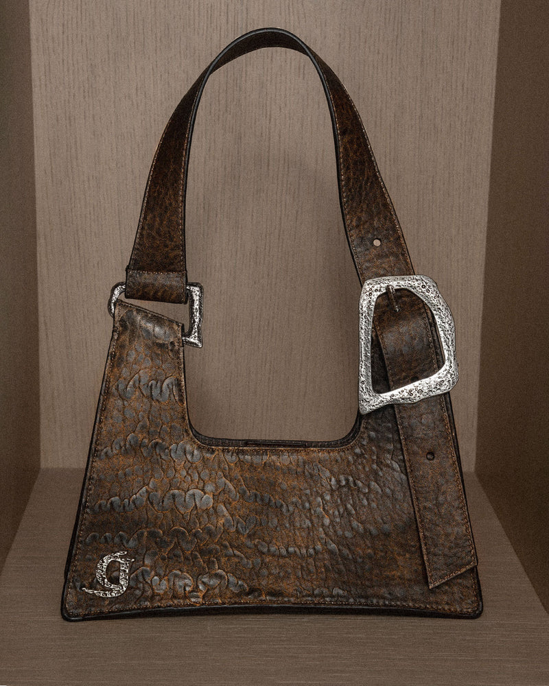 A still life photograph of a brown pebbled leather handbag with a statement buckle, set against a sleek, neural background.