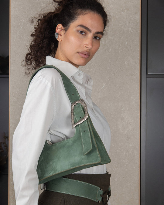 A stylish woman wearing a white shirt with a casual look, carrying a bag made of sage suede leather with silver hardware details. The bag has a soft, textured surface that complements her relaxed outfit.