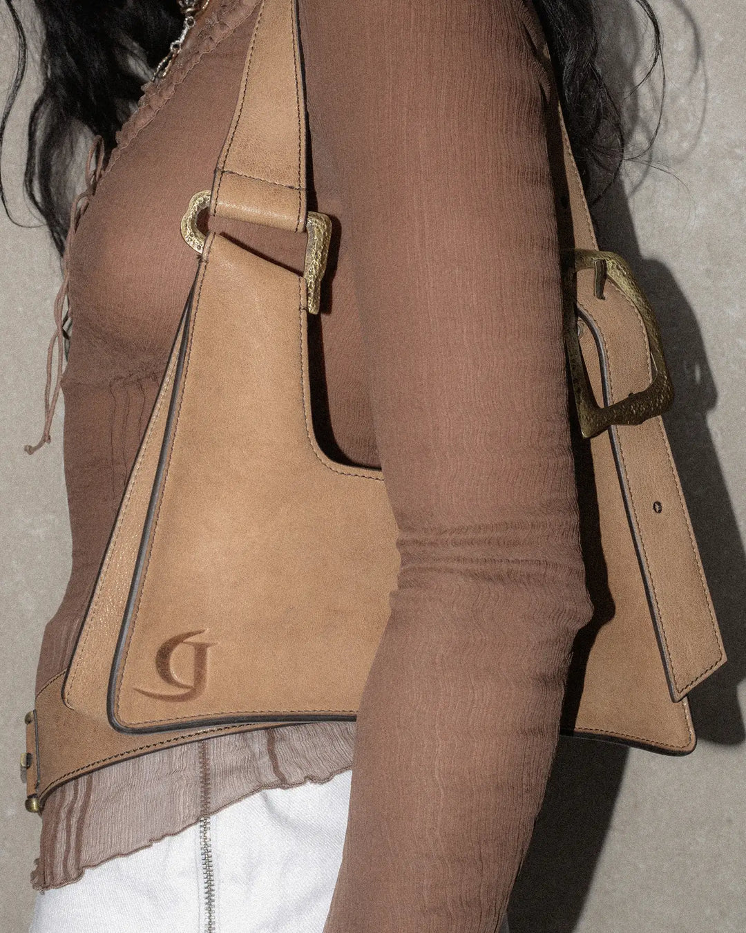 A model carrying a vintage tobacco-colored handbag slung over her shoulder. The bag features a textured, aged leather finish with a pressed logo visible in the left corner, adding a subtle branding detail. The overall look combines classic elegance with a touch of retro charm, highlighted by the worn-in, vintage texture of the bag.