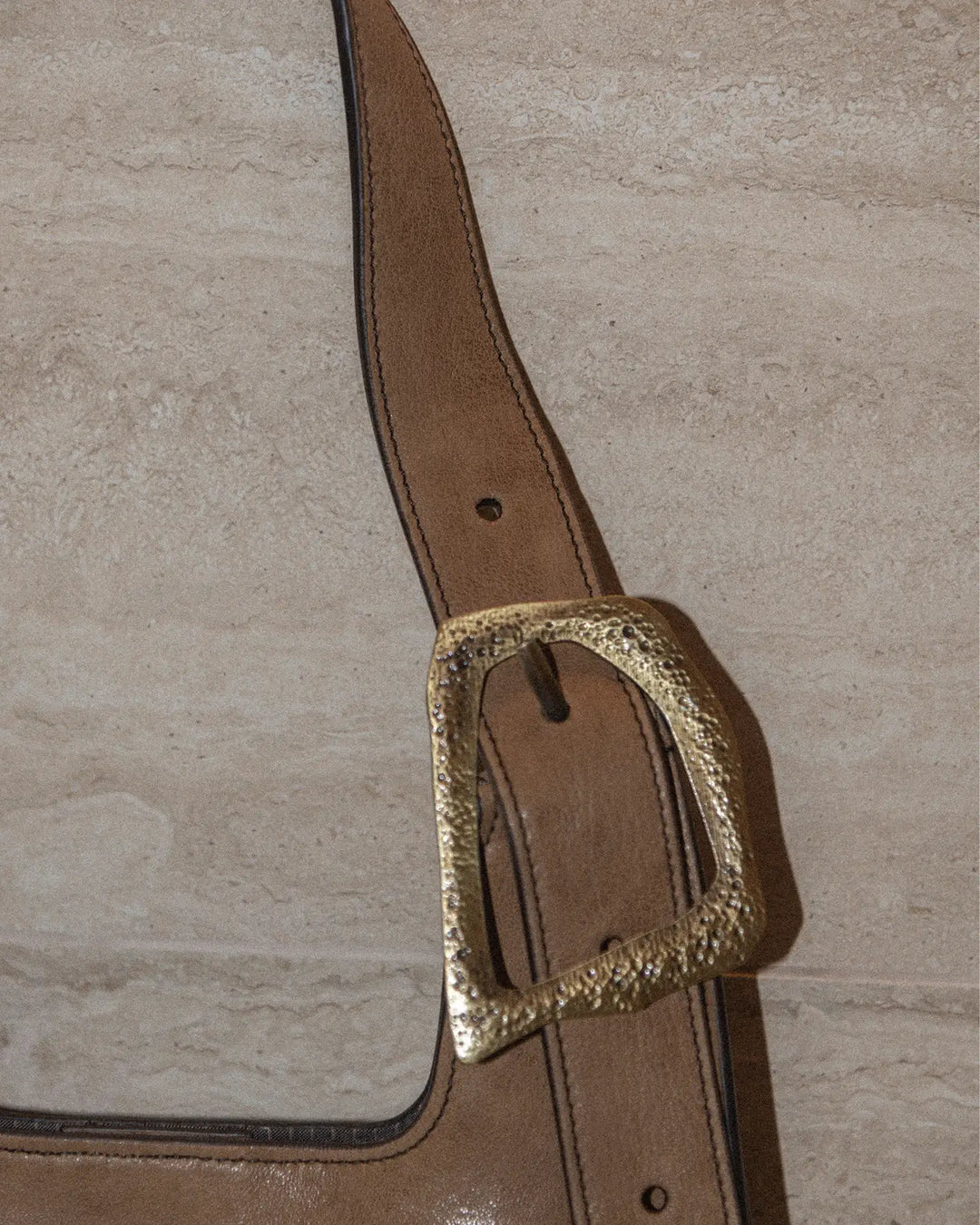 A close-up shot of a vintage tobacco-colored handbag featuring a distinctive buckle detail. The textured, aged leather showcases its timeless charm. The background is a luxurious marble surface, enhancing the sophisticated and elegant vibe of the scene.