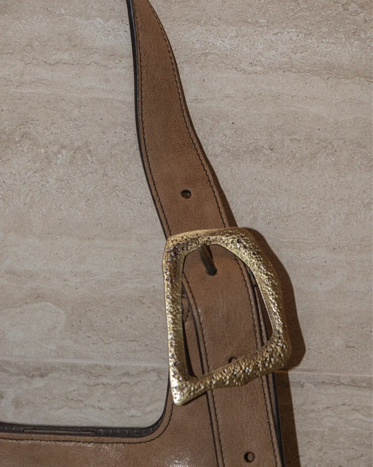 A close-up shot of a vintage tobacco-colored handbag featuring a distinctive buckle detail. The textured, aged leather showcases its timeless charm. The background is a luxurious marble surface, enhancing the sophisticated and elegant vibe of the scene.