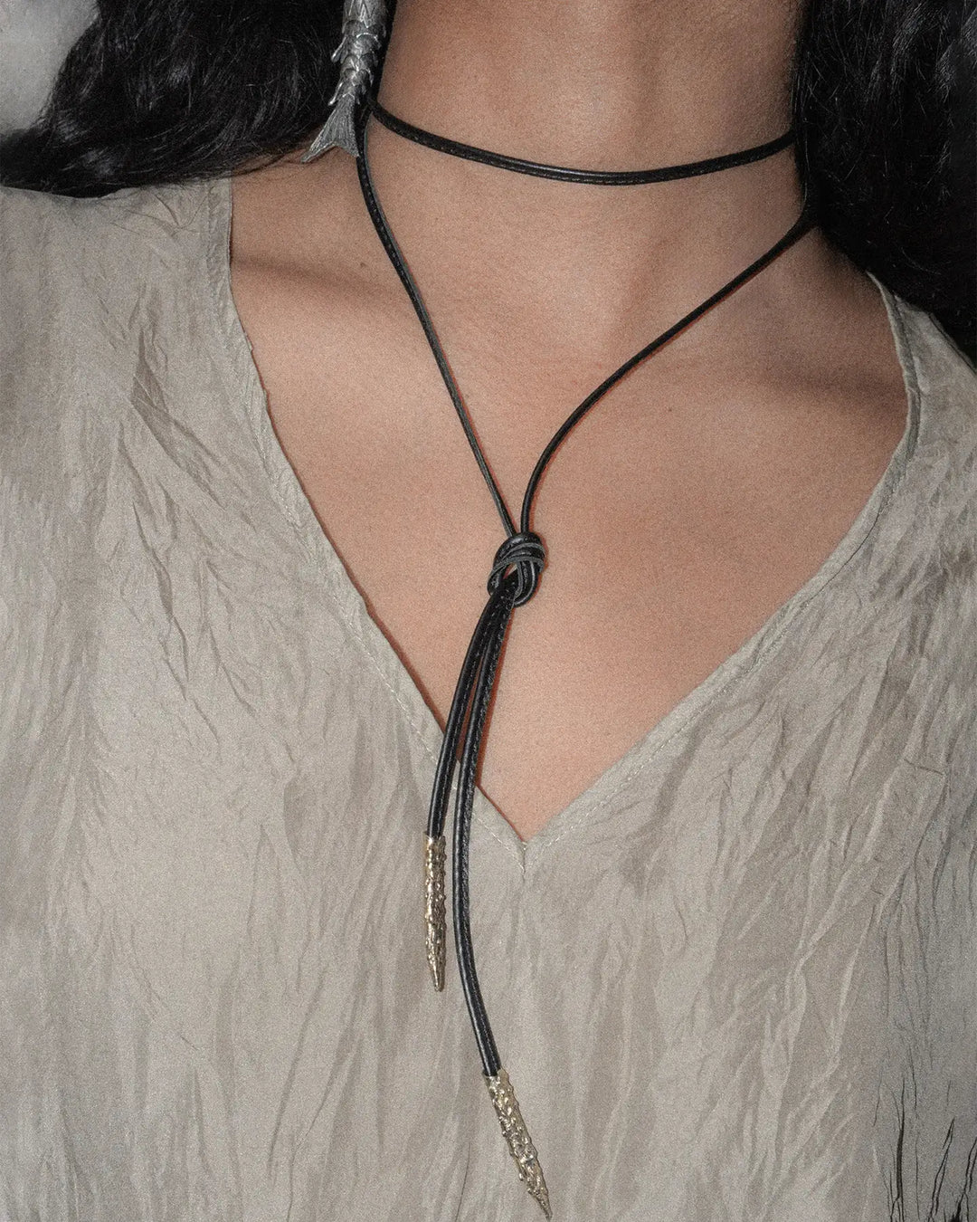 A close-up of the neck featuring a minimalist black leather ribbon styled twice around it. The ribbon is neatly wrapped, creating a layered, elegant look against the smooth skin. The subtle bronze details add a touch of sophistication, and the silk top provides a soft, luxurious backdrop.