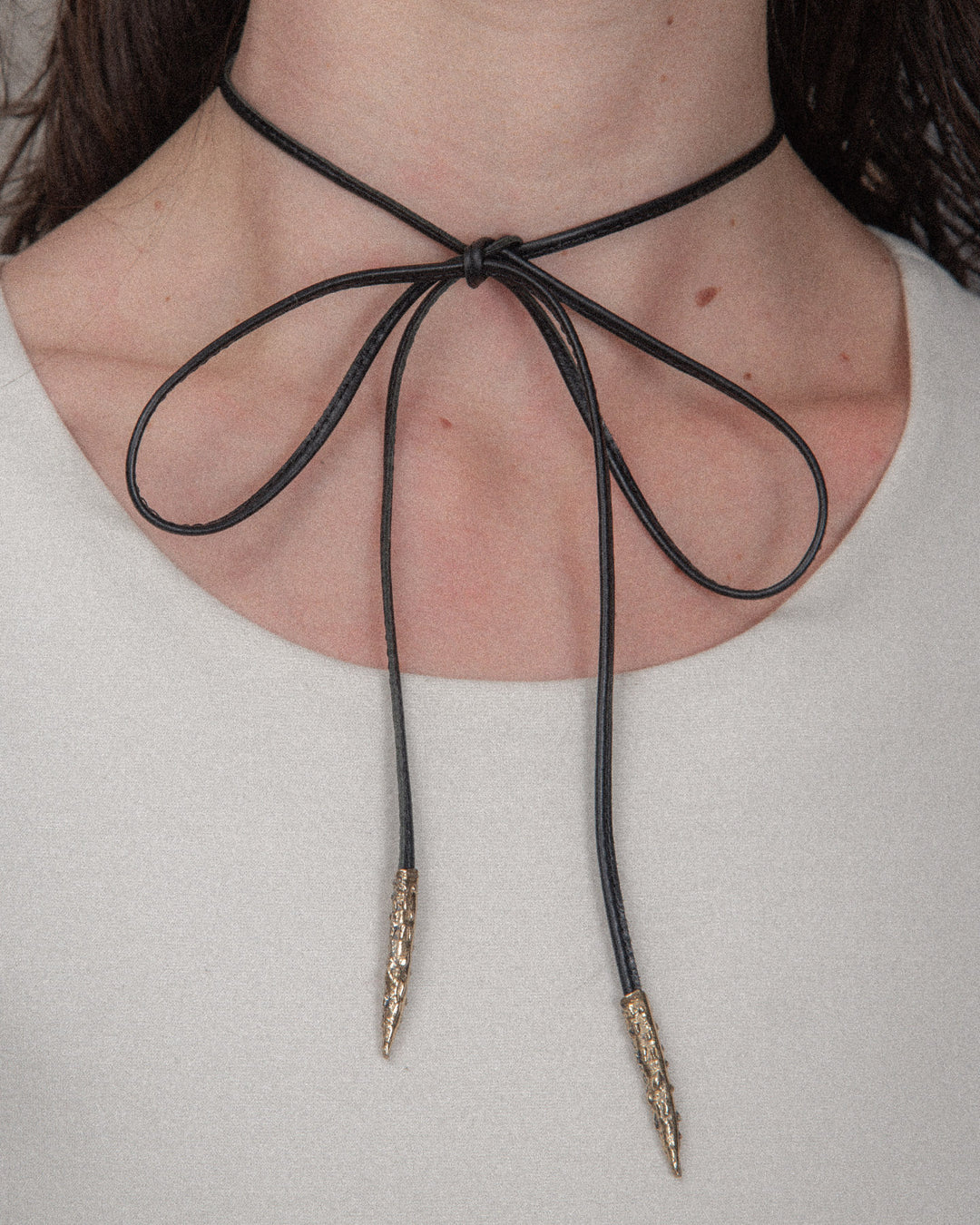 A large black leather ribbon with bronze details is styled as a bold bow on the white top. The ribbon is prominently placed at the neckline, creating a striking contrast against the white fabric. The oversized bow adds a touch of elegance and modern flair to the overall look.