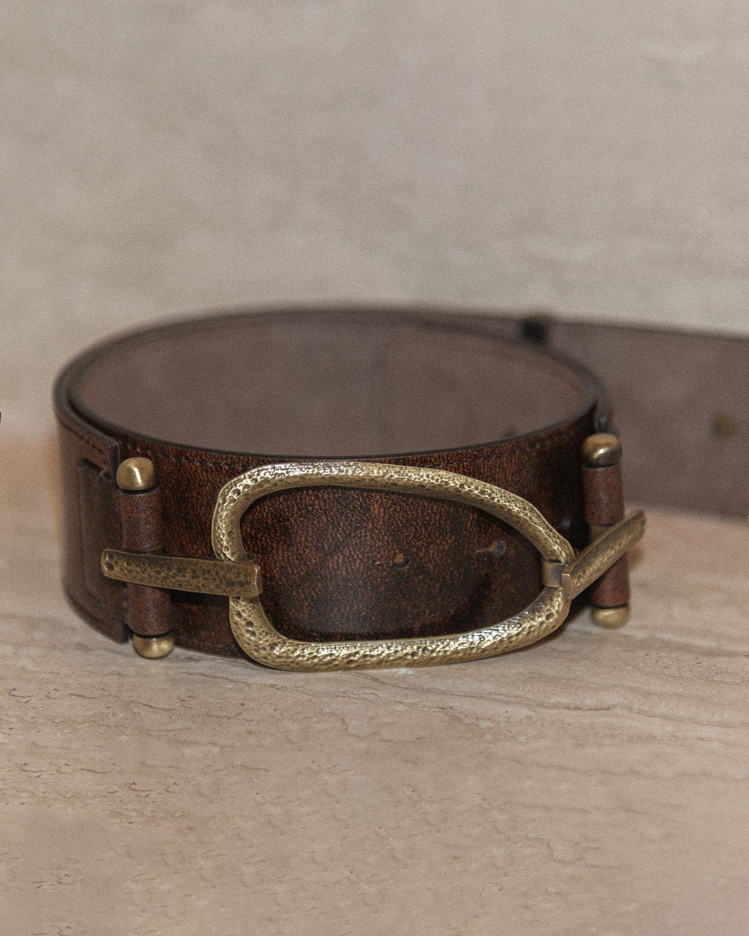 A still life of a belt with a chocolate brown strap and bronze hammered hardware, displayed against a neutral background. The image highlights the rich texture of the leather and the intricate hammered details of the hardware.