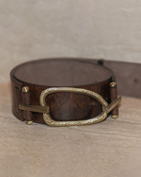 A still life of a belt with a chocolate brown strap and bronze hammered hardware, displayed against a neutral background. The image highlights the rich texture of the leather and the intricate hammered details of the hardware.