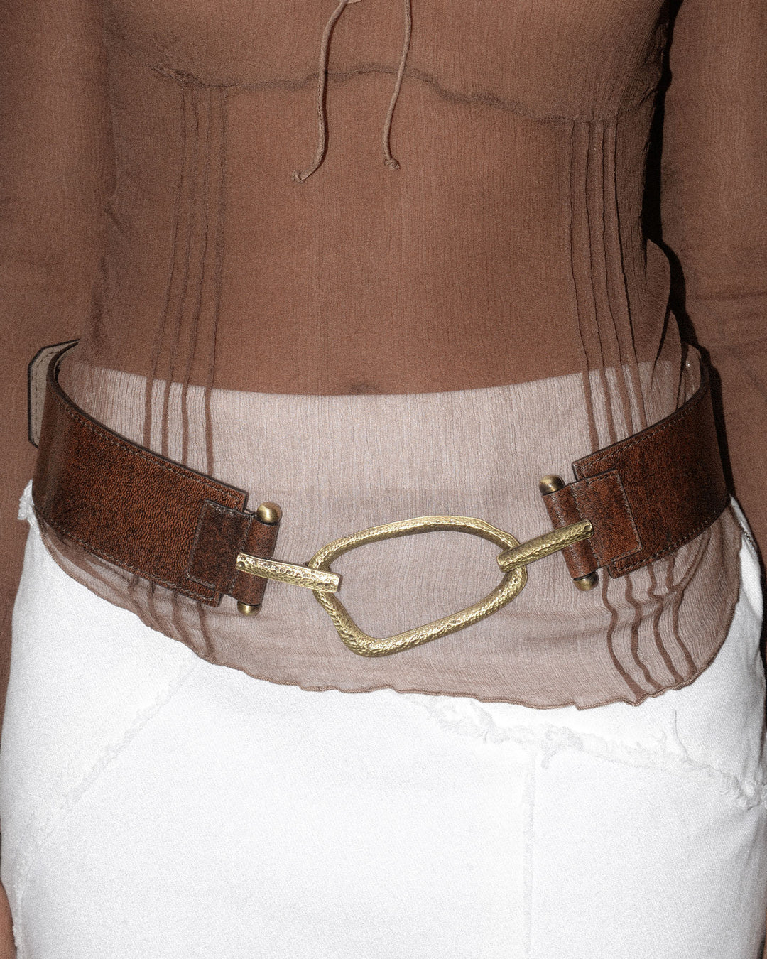 A woman wearing a white skirt and a brown top, showcasing a belt with a chocolate brown strap and bronze hammered hardware. The belt, styled on the hips, adds a chic and sophisticated touch to her stylish outfit.