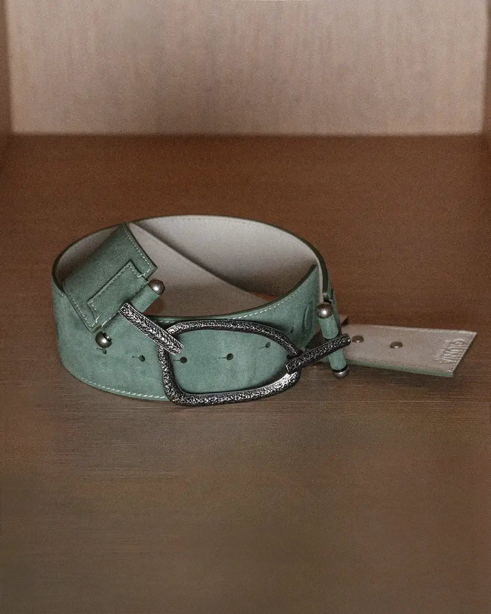 A still life photo of a sage suede belt with white stitching, laid flat on a wooden surface. The belt is positioned to show the backside detail