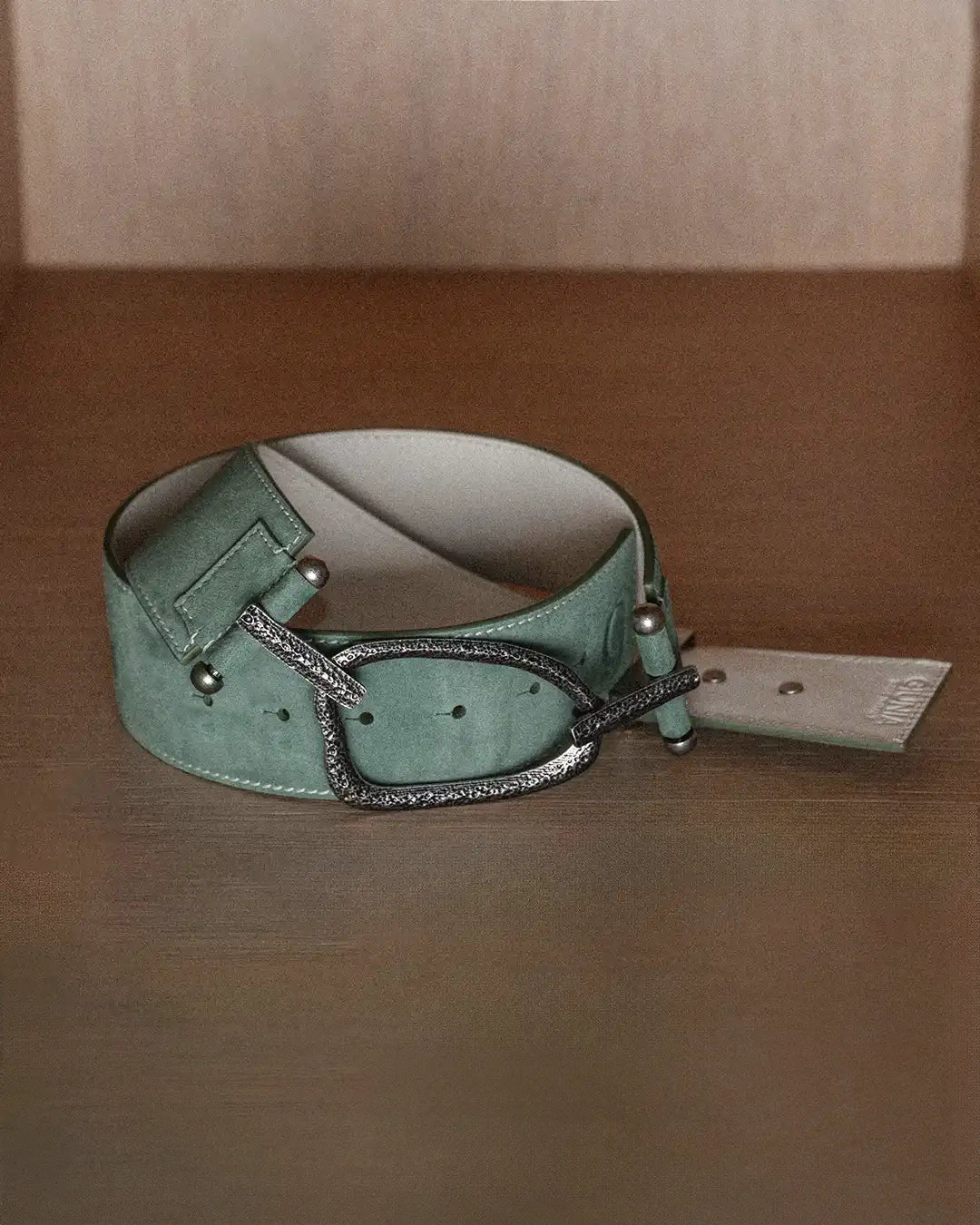 A still life photo of a sage suede belt with white stitching, laid flat on a wooden surface. The belt is positioned to show the backside detail