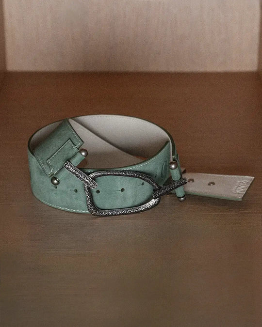A still life photo of a sage suede belt with white stitching, laid flat on a wooden surface. The belt is positioned to show the backside detail