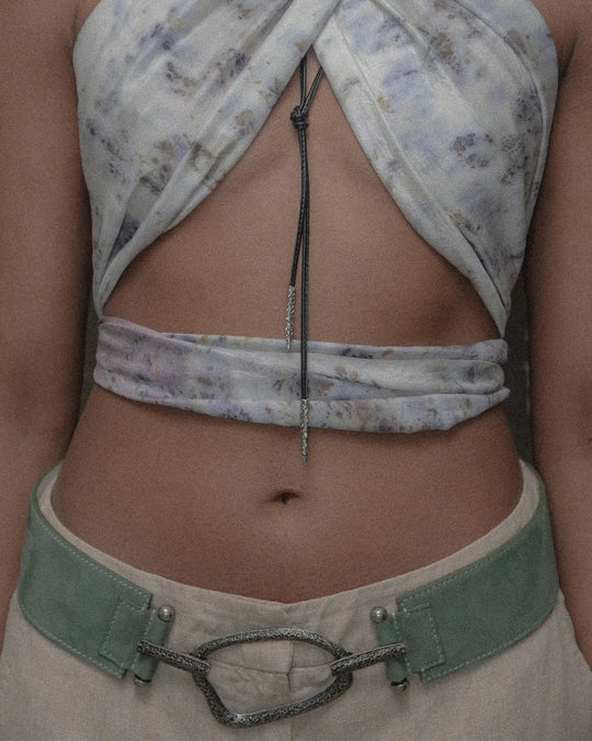 A woman wearing a sage suede belt with a statement silver detail, paired with beige linen pants and a silk top for a casual yet chic summer look. The view is from the front, emphasizing the stylish belt and its silver accent.