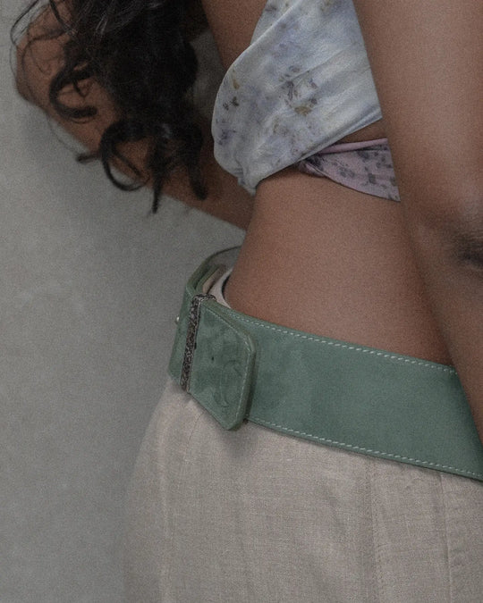A woman wearing a sage suede belt with a statement silver detail, paired with beige linen pants and a white contrast top with visible topstitching, for a casual yet chic summer look. The view is from the side, highlighting the stylish belt and the contrasting top stitches.