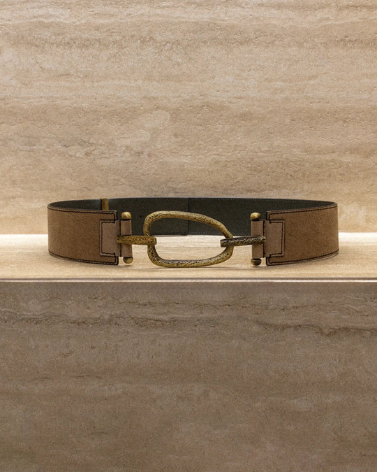 Still life of a brown vintage-finish belt with a hammered eye-shaped metal buckle, displayed on a marble surface.
