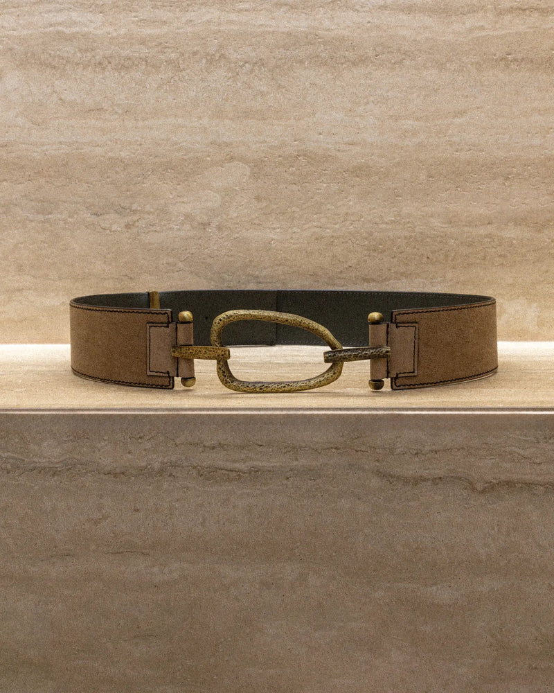 Still life of a brown vintage-finish belt with a hammered eye-shaped metal buckle, displayed on a marble surface.