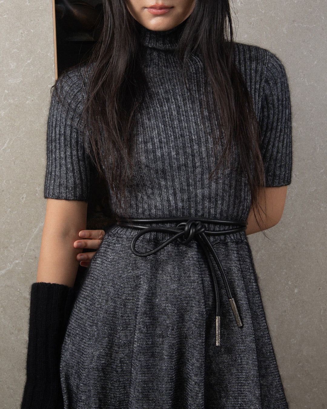 Front view of a tubular black leather belt with silver metal details, featuring a stunning knot that adds a touch of elegance and texture. The belt sits at the waist, complementing a knitwear dress with a sophisticated and stylish look