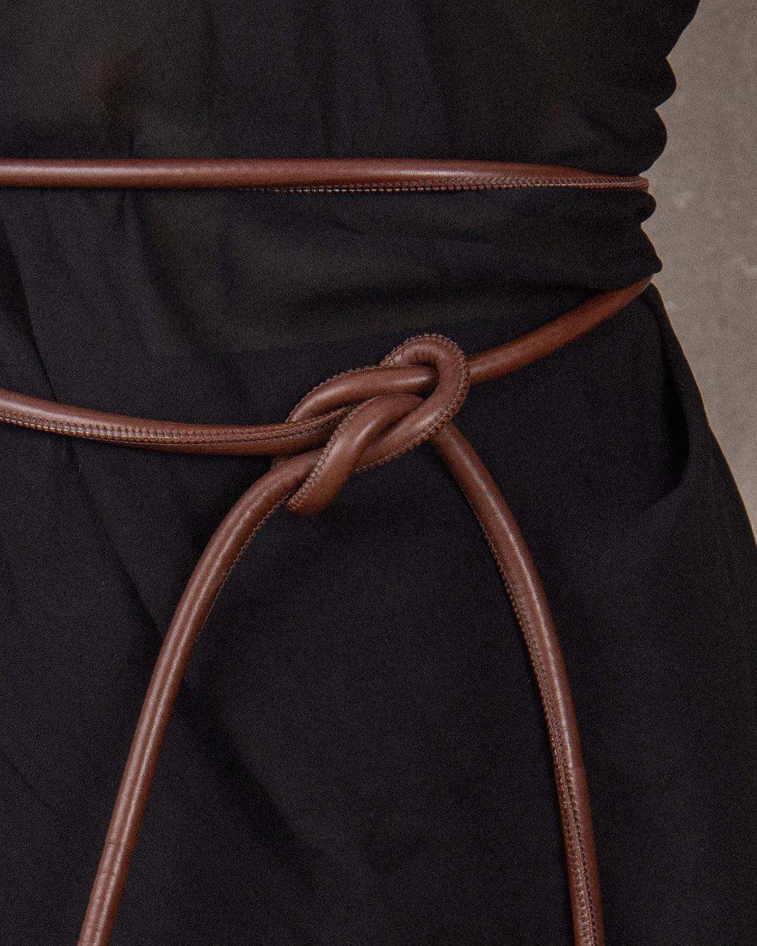 Close-up of a tubular leather belt with textured bronze metal details, tied in an elegant knot on a black sheet top, showcasing its versatile design and craftsmanship