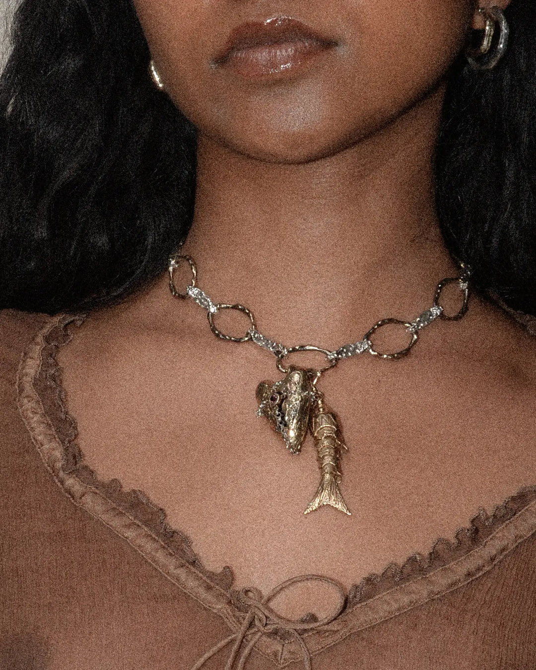 A close-up of a bronze fish-shaped charm with textured details and gemstone eyes, glinting softly on a delicate chain worn by a woman in a luxurious brown silk blouse.