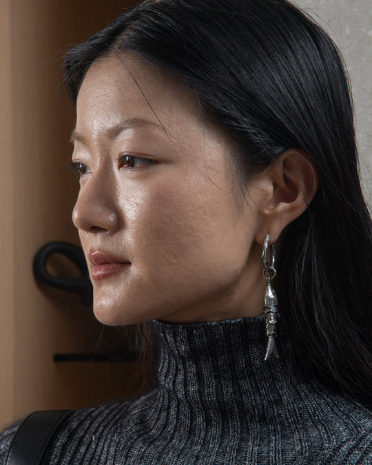The silver fish-shaped charm with textured scales and peridot gemstone eyes is dangling from an earring, highlighting its intricate design and craftsmanship.